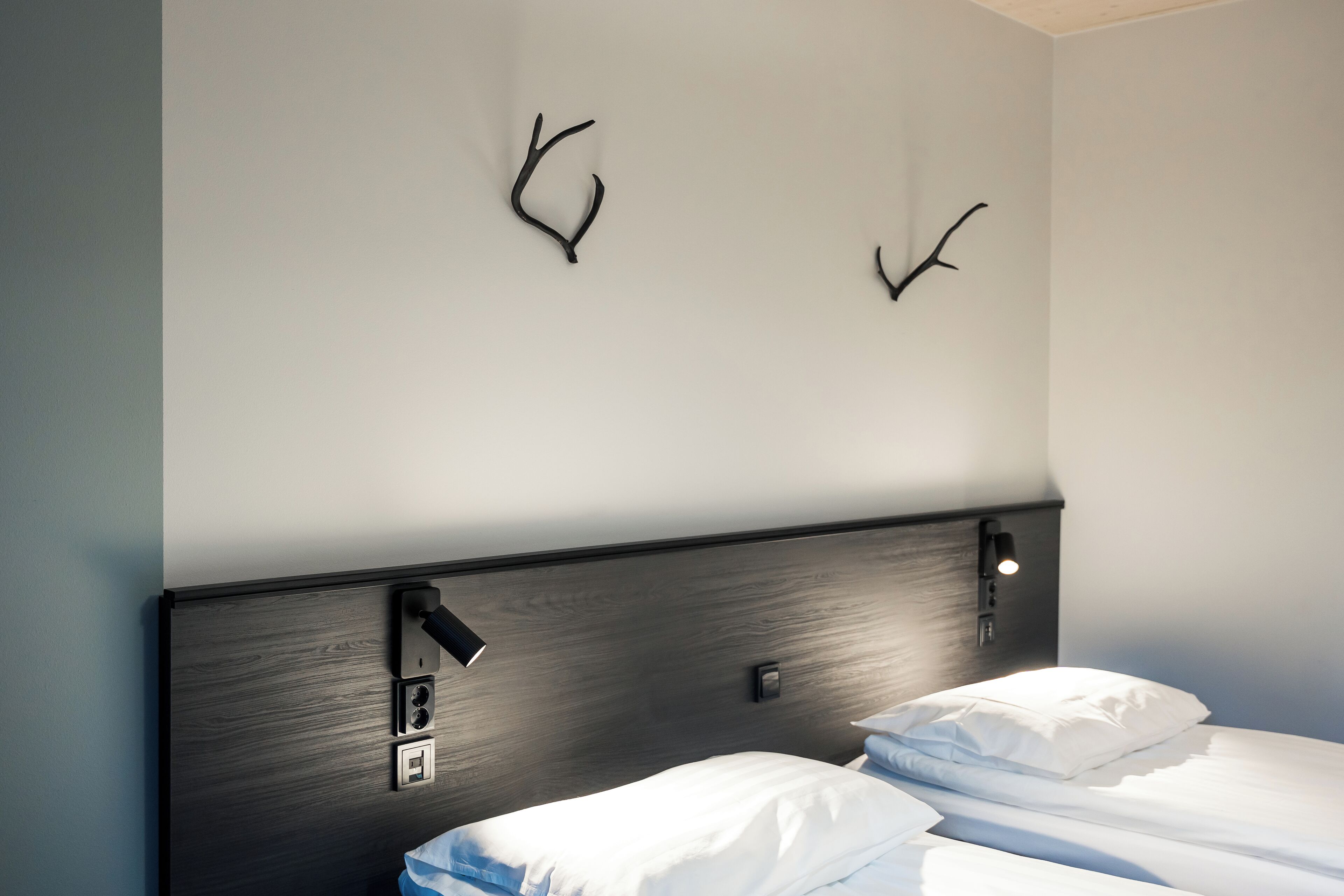 A hotel room with a dark wood headboard, two wall-mounted hooks, and a bedside table with a lamp.