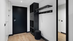 Un pasillo con una puerta negra, suelo de madera, un cesto de basura negro y un perchero negro con ganchos.