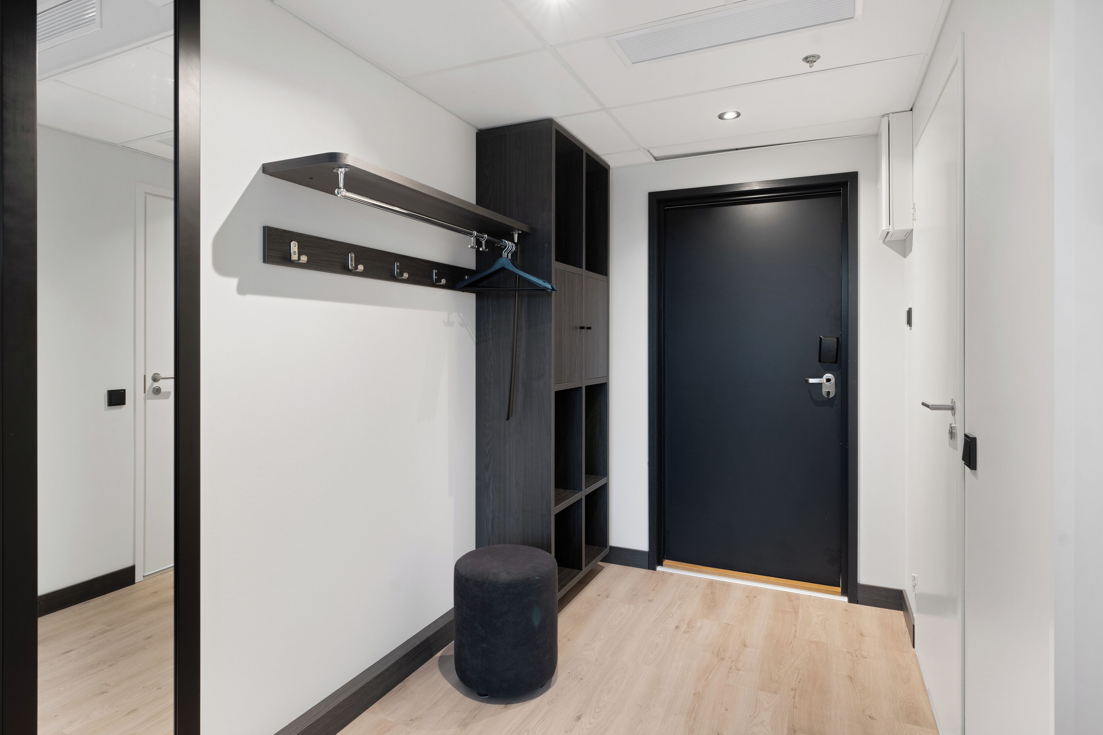 Et moderne hotelværelse med trægulv, dør i sort, knagerække og lille sort fodskammel.