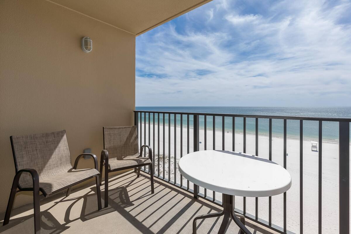 Un balcon avec une table blanche et deux chaises en osier, donnant sur une plage protégée par une rambarde en métal.