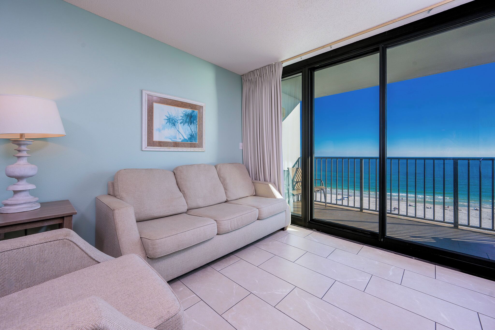 Un balcone con vista sulla spiaggia, un divano color beige, una lampada e un quadro appeso al muro.