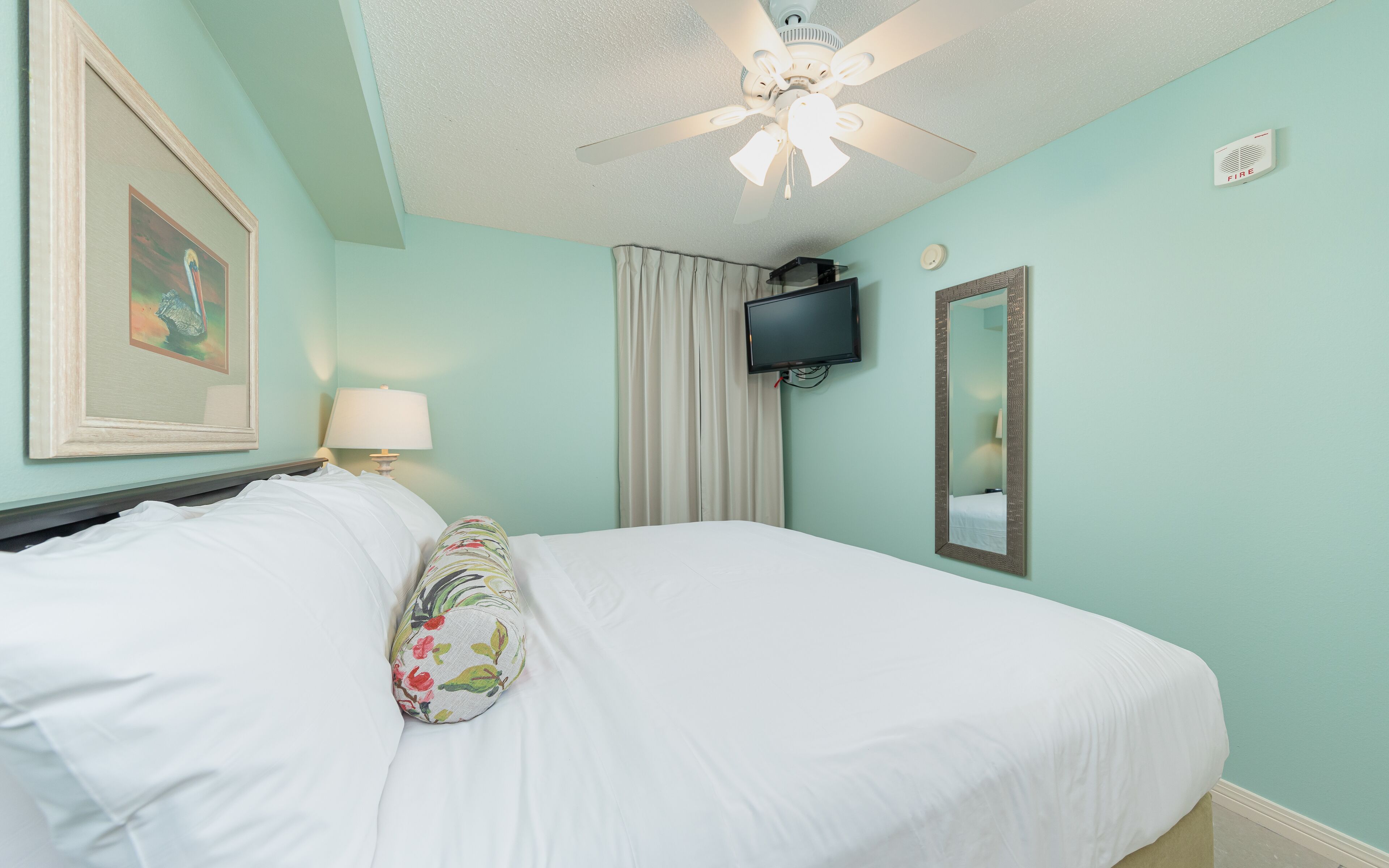 A neatly made bed with white linens and a floral pillow, a ceiling fan, a mounted TV, a fire alarm, and a framed picture on the wall.