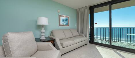 A room with a sofa, armchair, and a table with a lamp. There's a painting on the wall and a view of the ocean through a balcony.