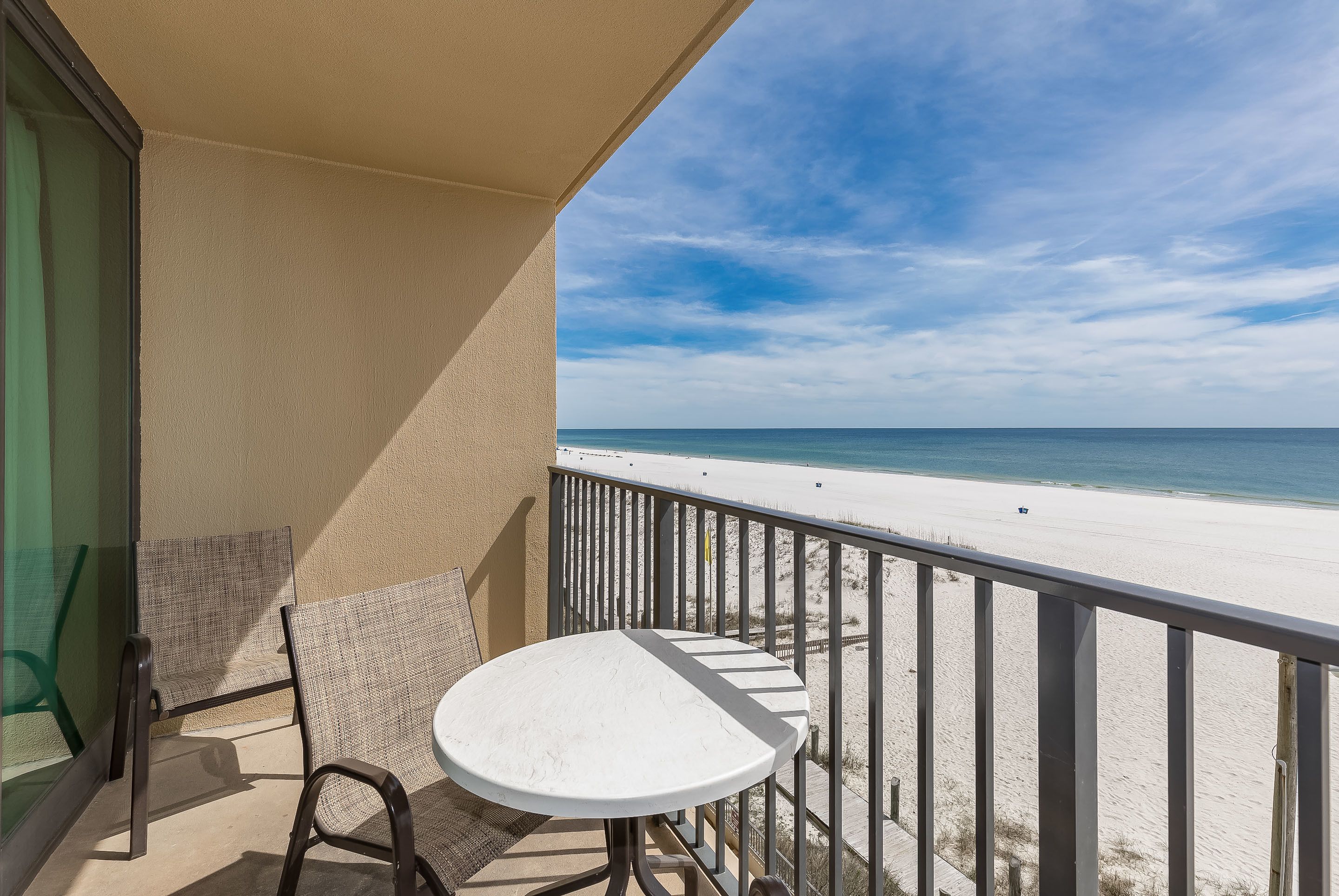 A balcony with a white table and two wicker chairs, overlooking a beach with a clear sky.