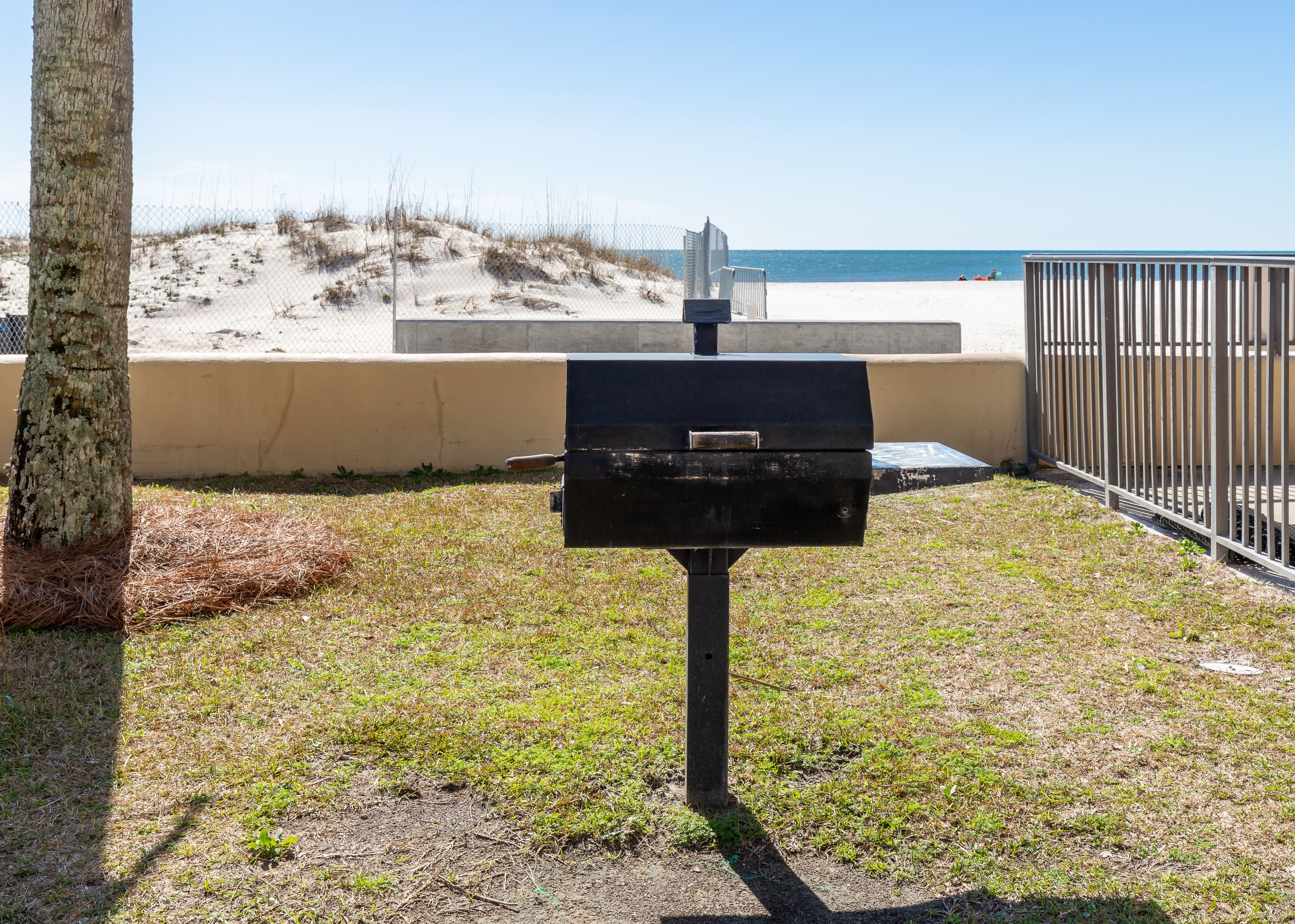 A barbecue grill in a grassy area with a beach and a fence in the background.