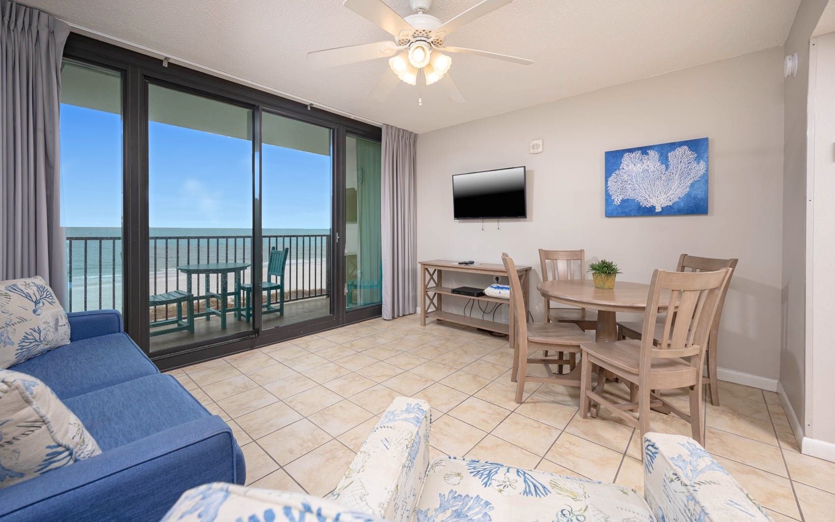 Una habitación con un sofá azul, una mesa de comedor con sillas, un televisor en la pared y vista a la playa a través de una puerta corrediza de vidrio.