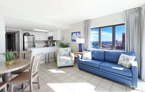Una sala moderna con un sofá azul, un comedor con una mesa y sillas de madera, y una cocina con armarios y electrodomésticos blancos.