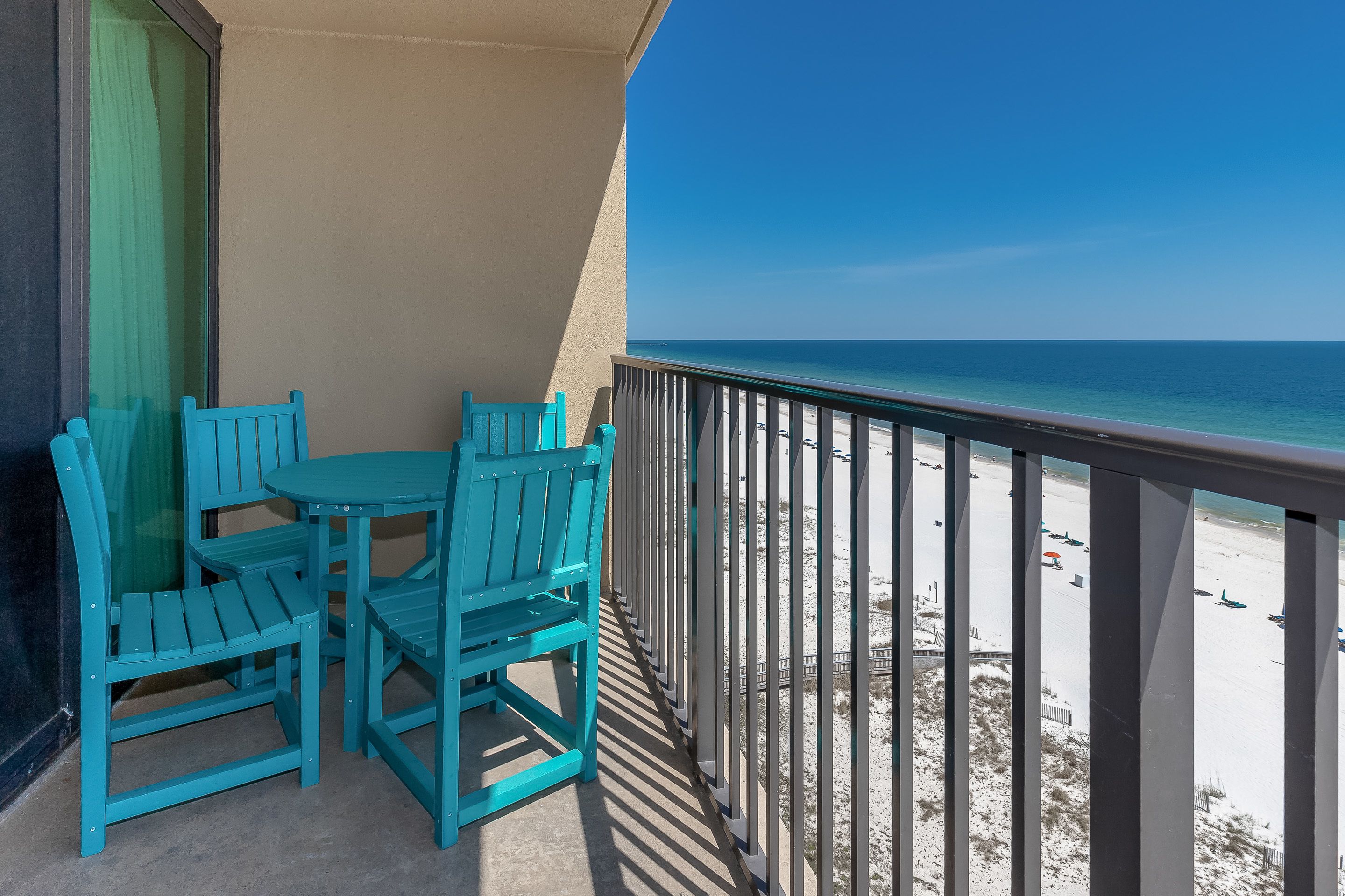 A balcony with turquoise chairs and a table, overlooking a beach with a clear blue sky.