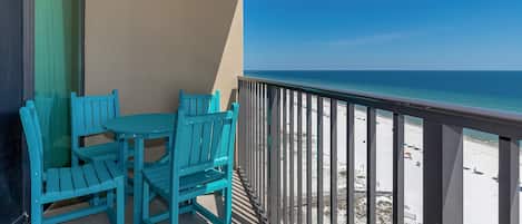 A balcony with turquoise chairs and a table, overlooking a beach with a clear blue sky.