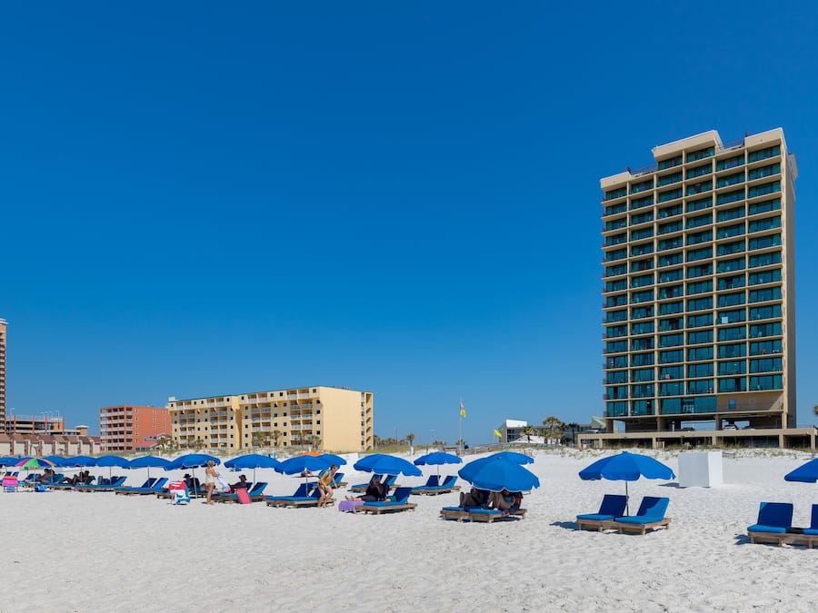 A beach with rows of sun loungers and blue umbrellas, a tall hotel building, and other buildings in the background.
