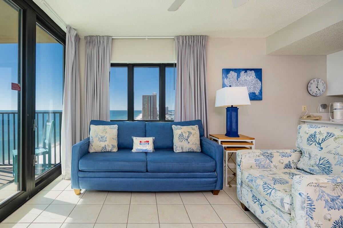 A balcony with a glass door leading to a terrace, a blue sofa with decorative pillows, a patterned armchair, a lamp, and a wall clock.