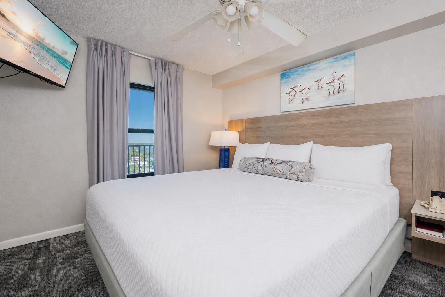 Ein Hotelzimmer mit einem großen Bett, einem Deckenventilator, einem Gemälde, einem Fenster mit Vorhängen sowie einem Nachttisch mit Lampe und Telefon.