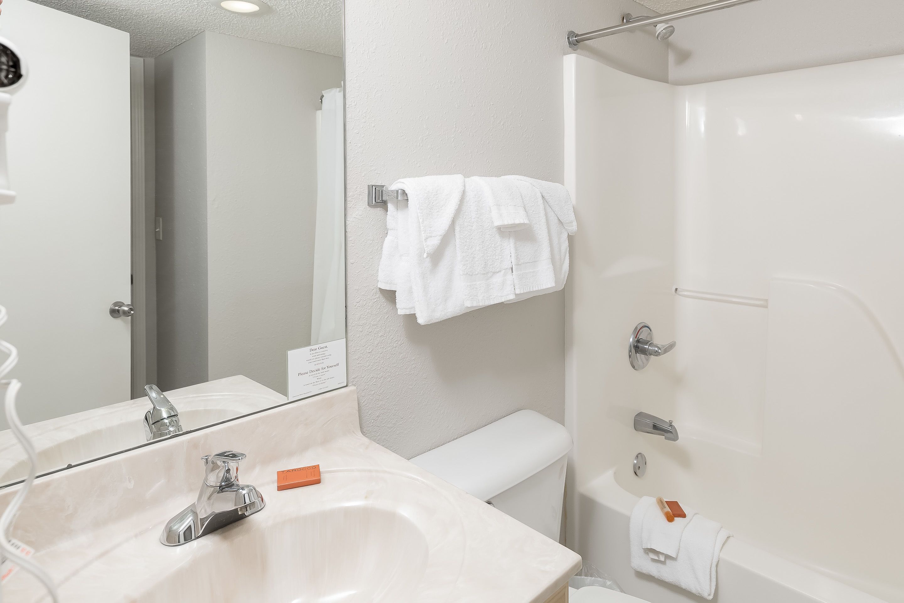 A bathroom with a white sink, mirror, and bathtub. There are towels hanging on the wall and a sign above the mirror.