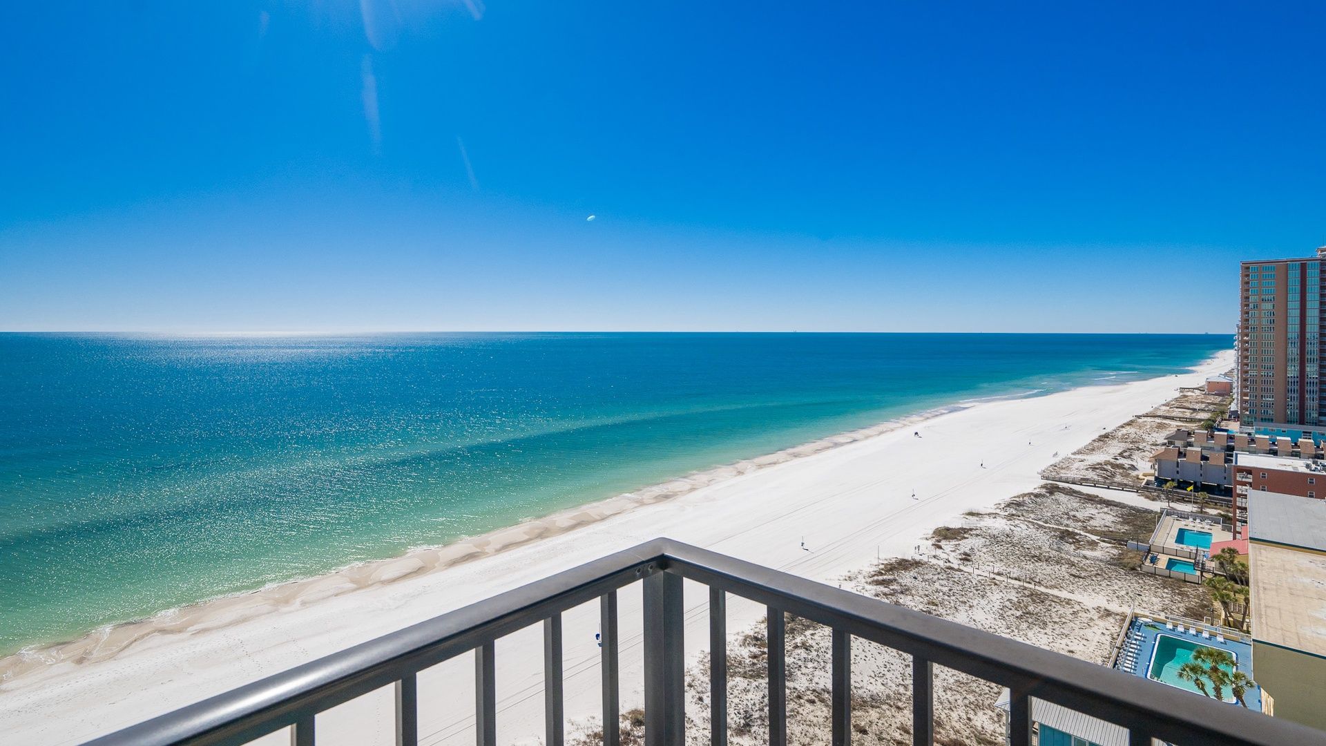 A beach view from a balcony with a clear sky, a sandy beach, and a high-rise building with a pool.