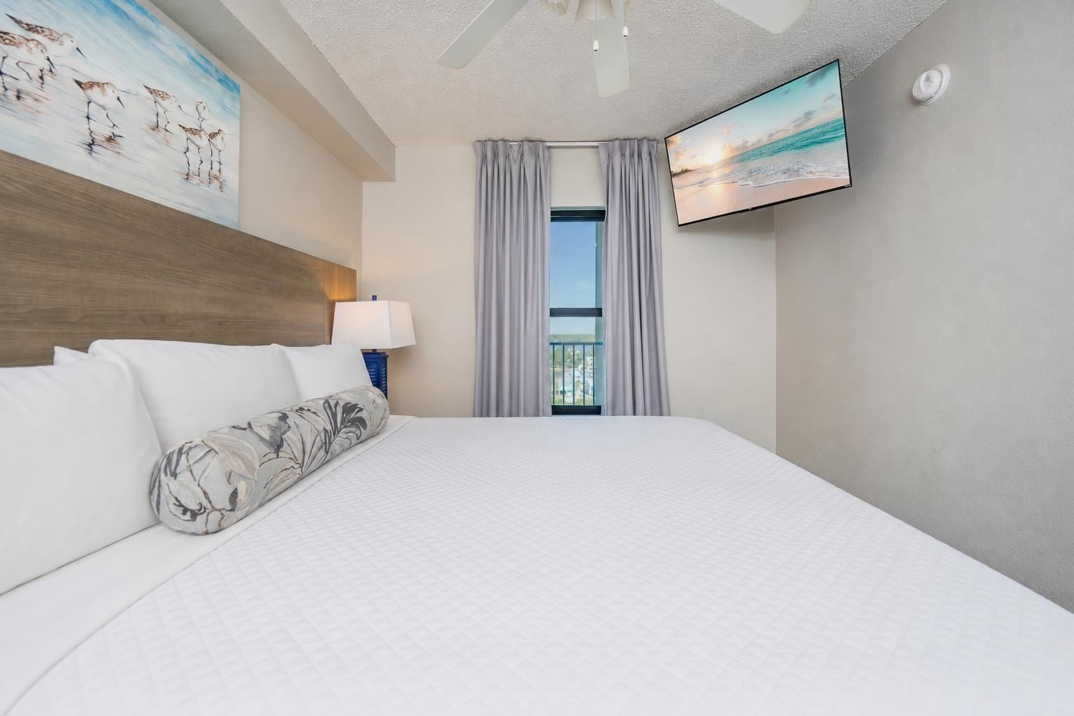 A hotel room with a large bed, a flat-screen TV, a ceiling fan, and a window with curtains.