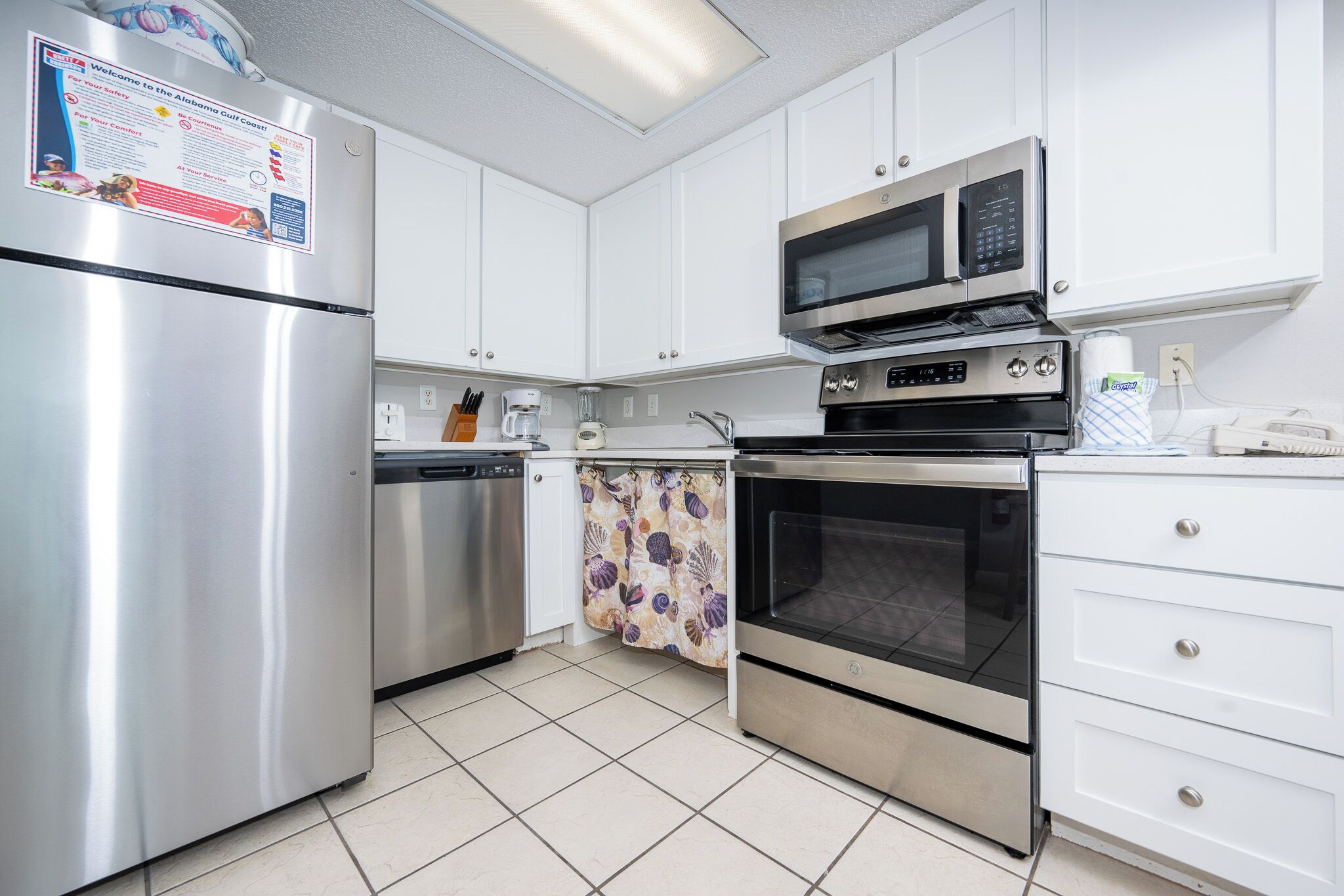 A kitchen with white cabinets, stainless steel appliances including a refrigerator, microwave, and oven, a dishwasher, and a patterned curtain.