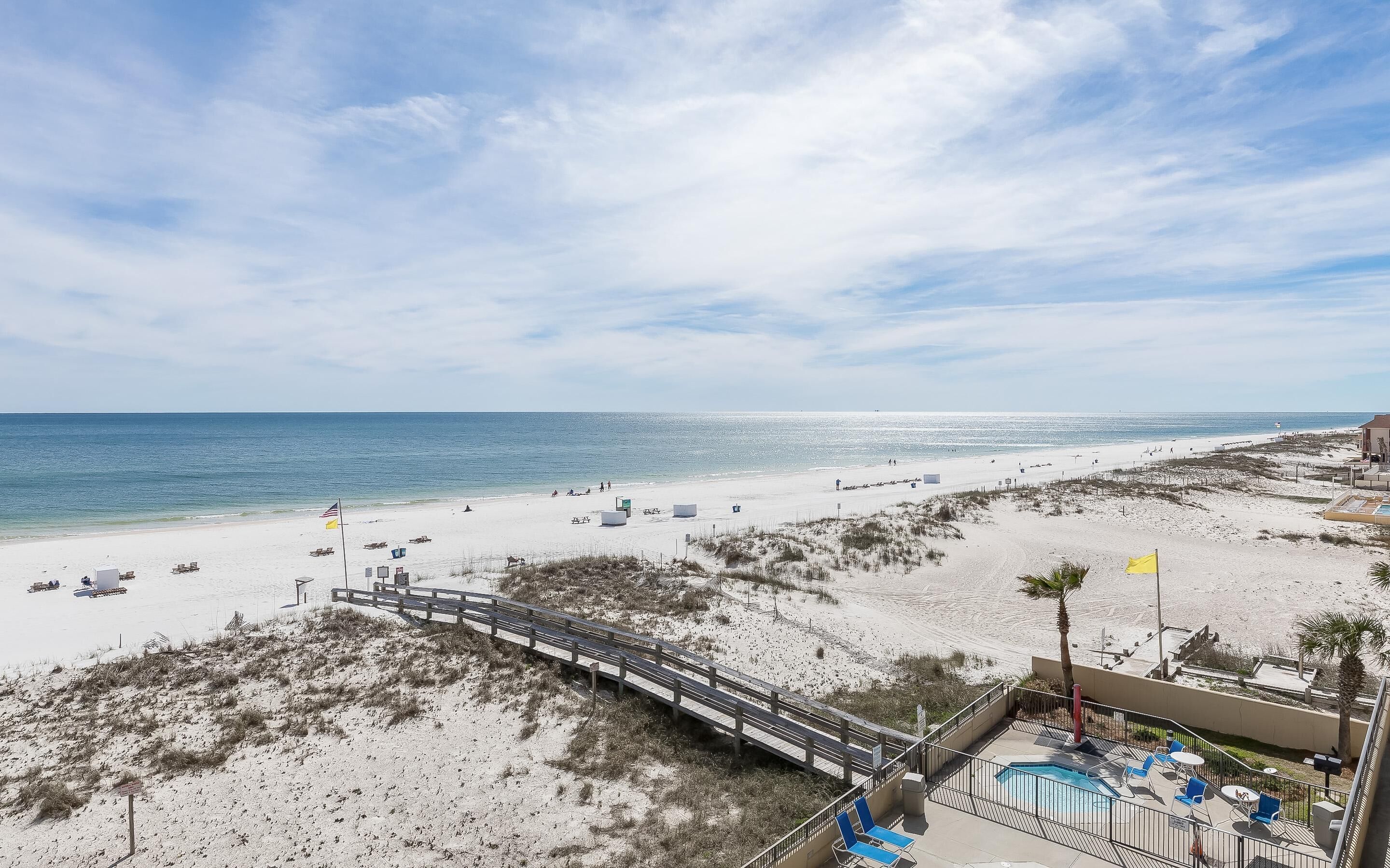 A beach with a wooden boardwalk leading to a pool area with lounge chairs.