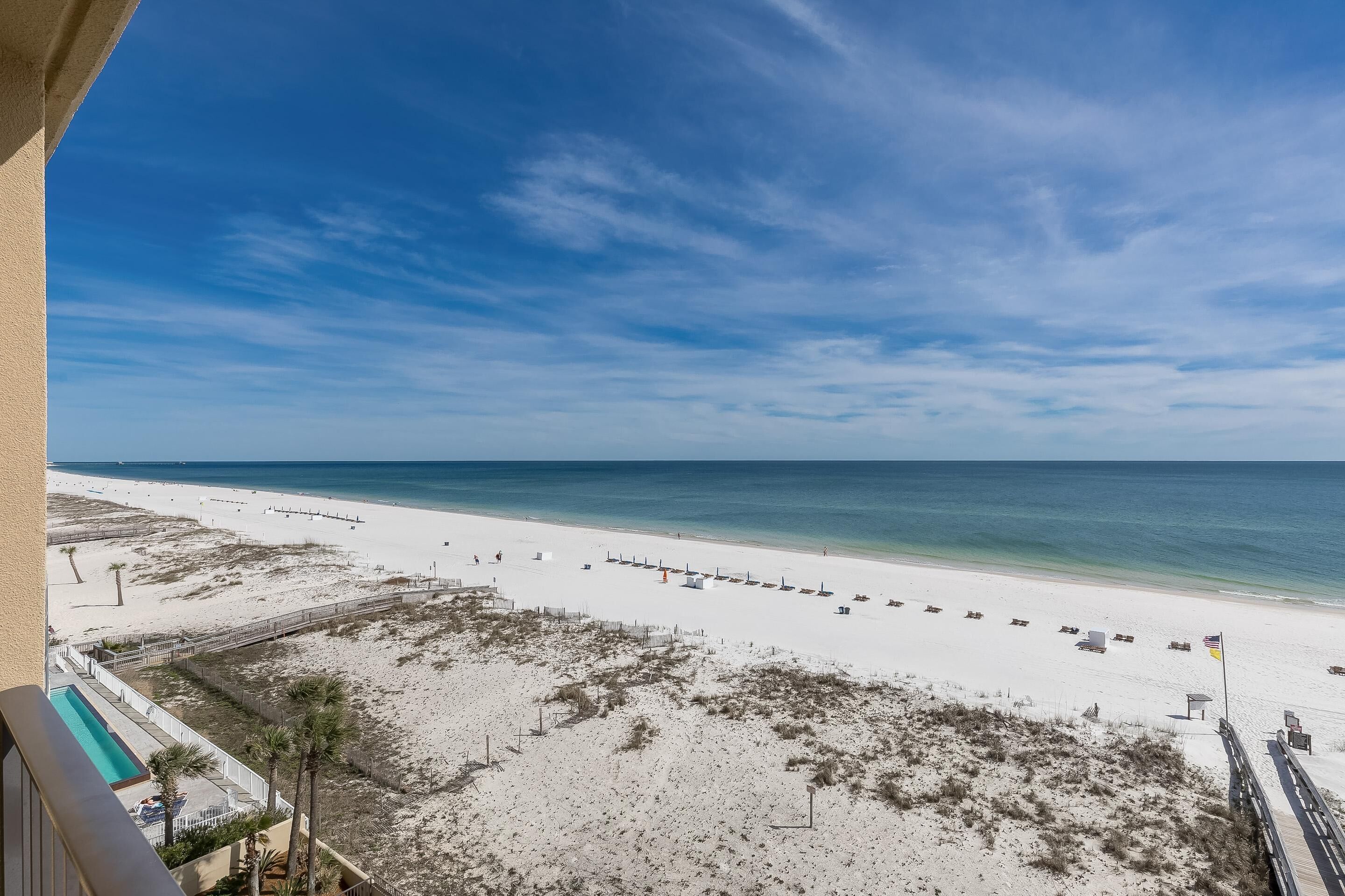A beach view from a balcony with a pool, beach chairs, and a flag.