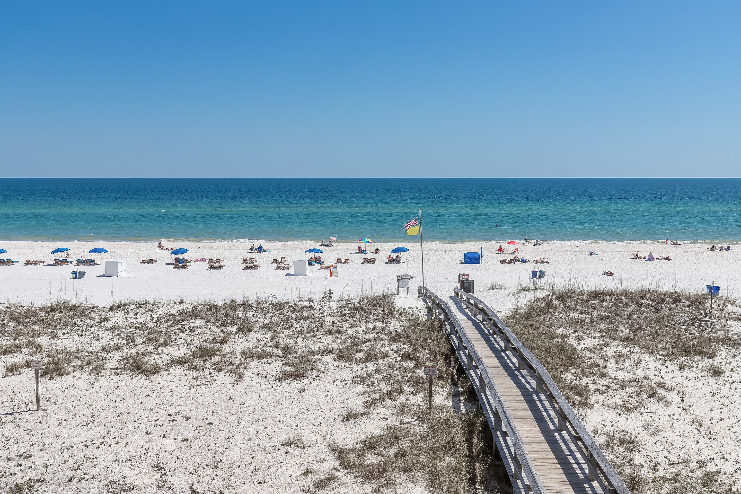 A beach with a wooden boardwalk leading to the water, white sand, and a flag.