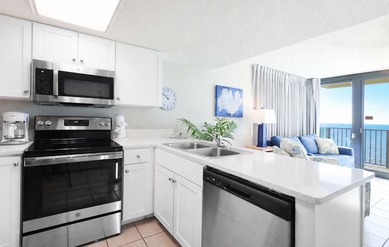 A modern kitchen with a dishwasher, oven, and microwave. There is a dining area with a blue sofa and a view of the sea.