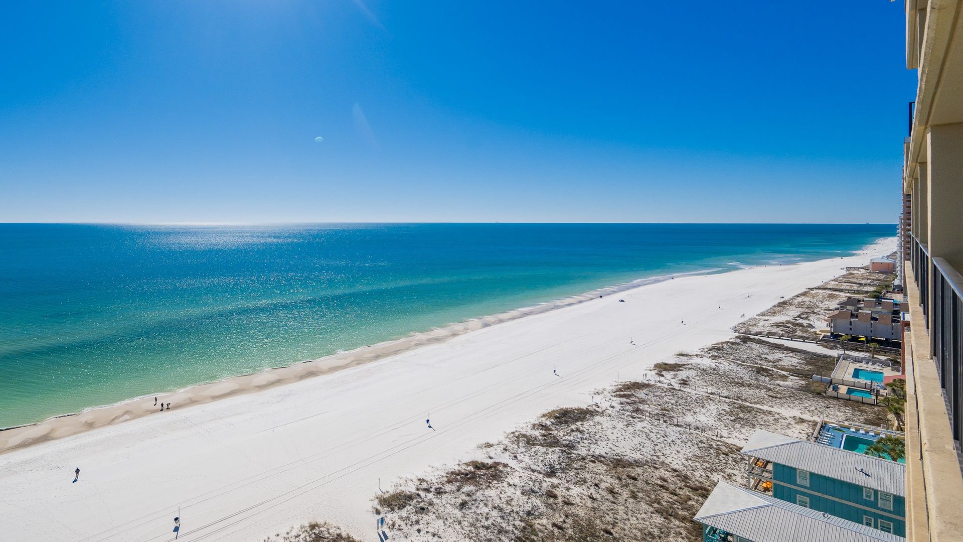 Blick auf einen Strand mit blauem Himmel, sandigem Ufer und einem Gebäude mit Balkon, das auf den Strand blickt.