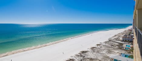 Blick auf einen Strand mit blauem Himmel, sandigem Ufer und einem Gebäude mit Balkon, das auf den Strand blickt.