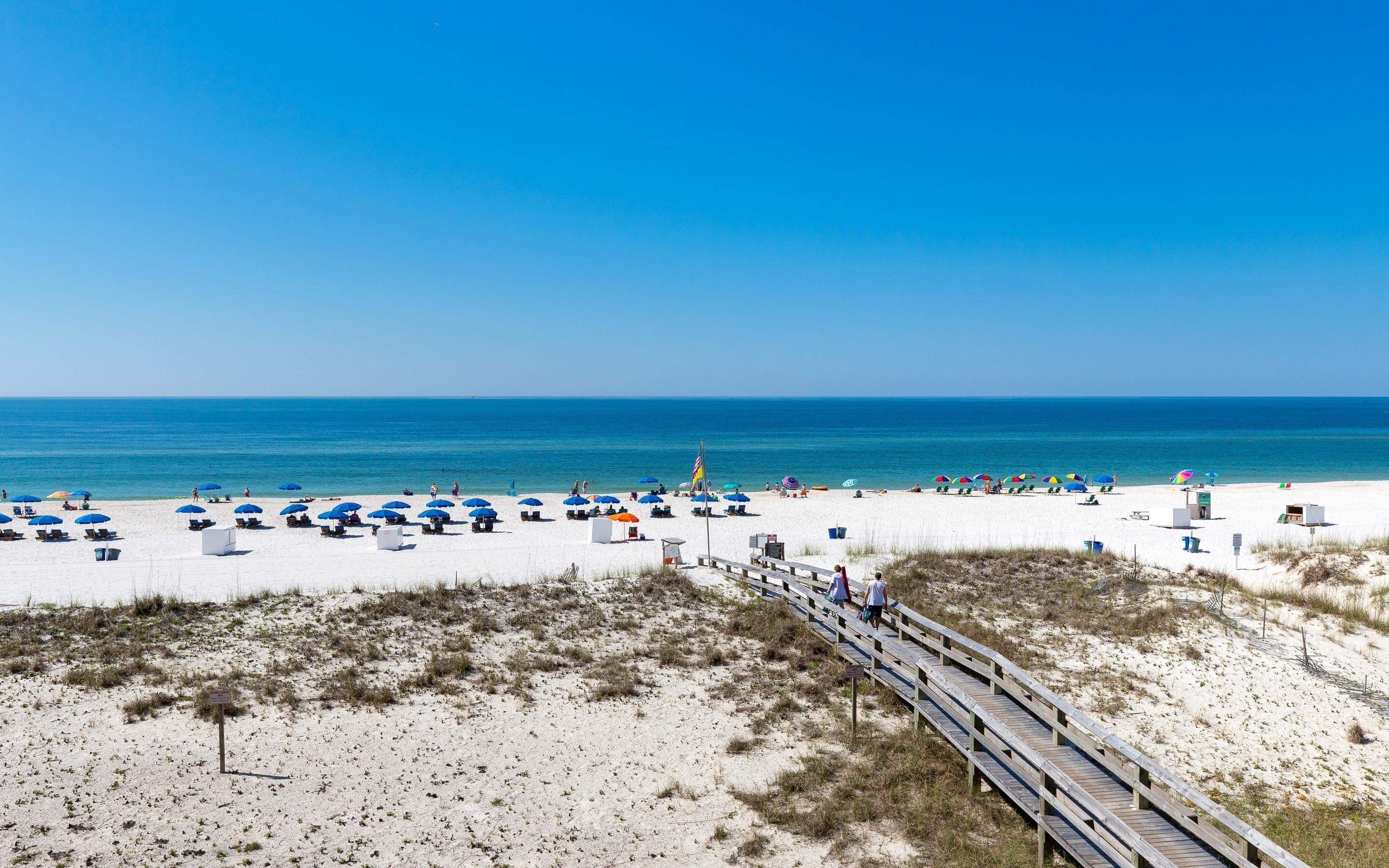 A beach with white sand, a wooden boardwalk, and blue umbrellas.