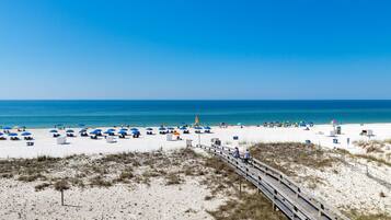 A beach with white sand, a wooden boardwalk, and blue umbrellas.