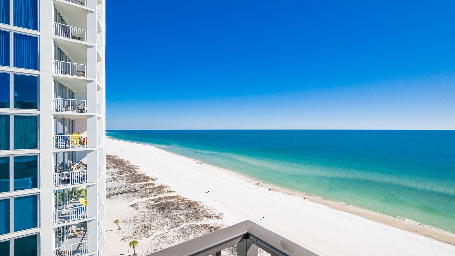 A beachfront hotel with a balcony overlooking a sandy beach and clear blue waters.