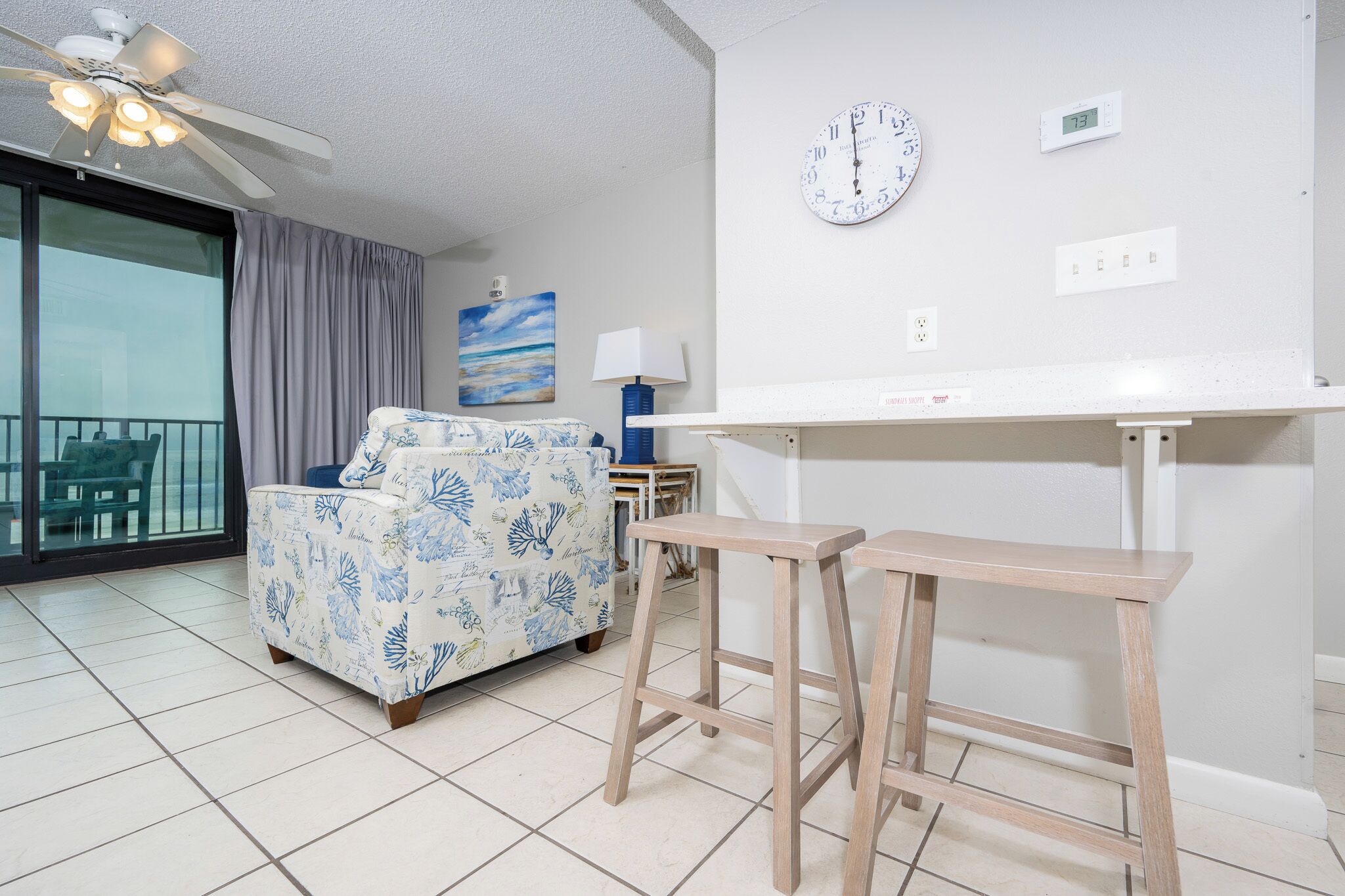 A room with a bed, a ceiling fan, a clock, and a digital thermostat.