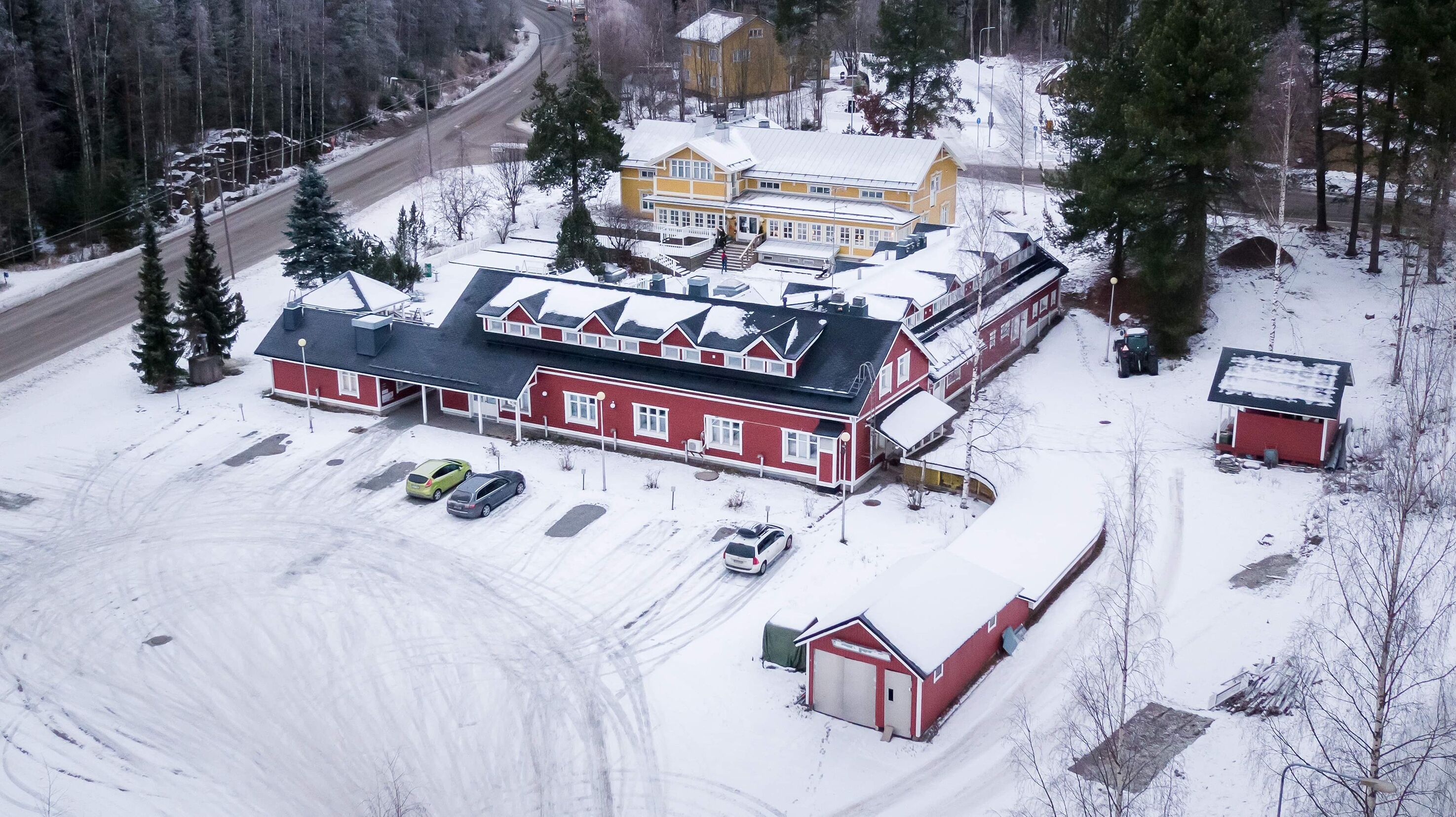 Un edifici gran vermell amb zona de pàrquing coberta, un edifici vermell més petit i un edifici groc en un paisatge nevós.