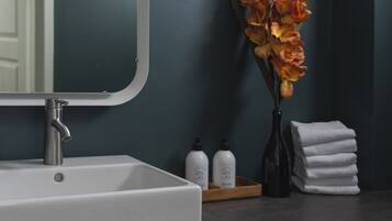 Une salle de bain avec un lavabo blanc, un miroir, un vase contenant des fleurs oranges et une pile de serviettes blanches.
