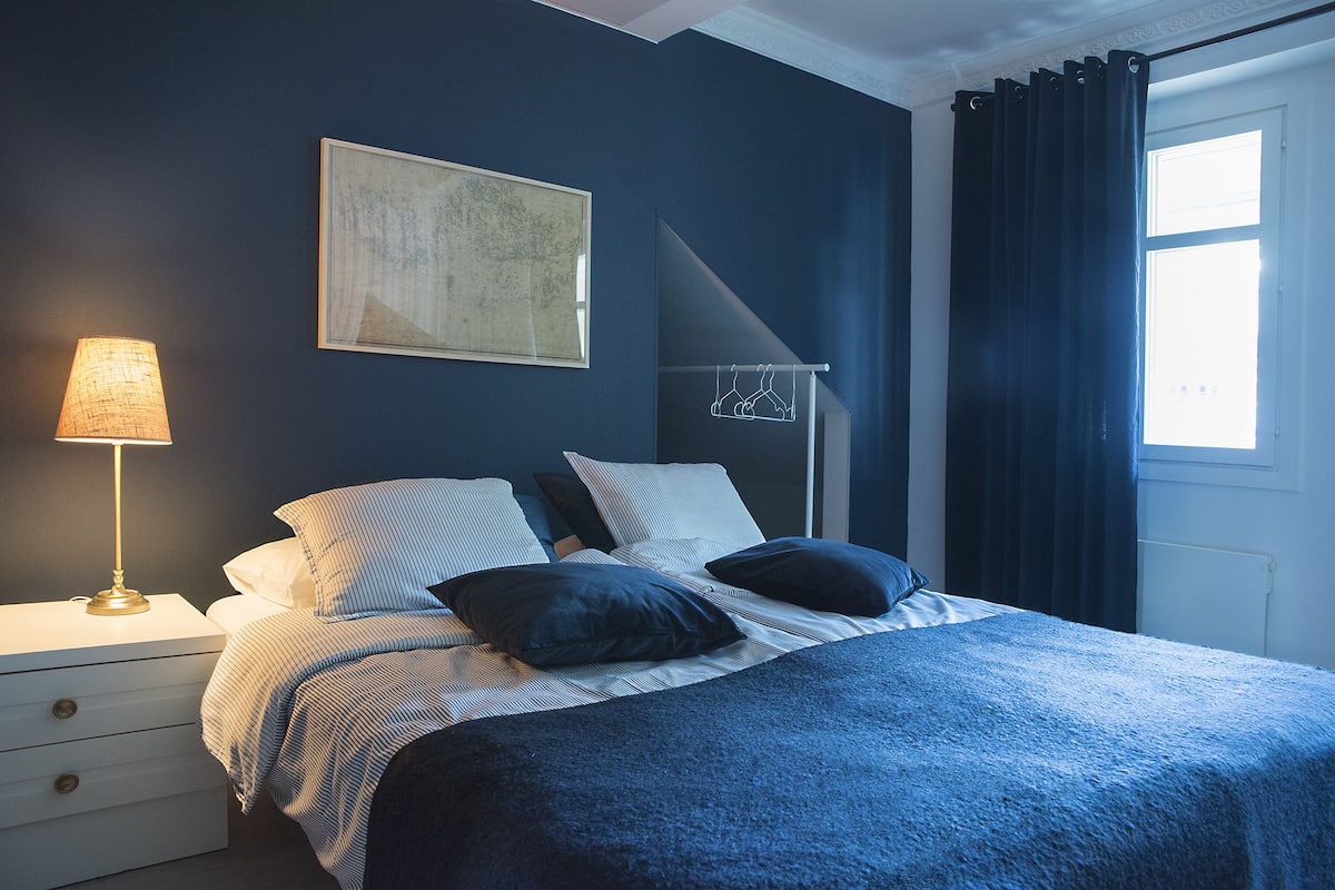 A bedroom with a bed, a nightstand, a lamp, a painting, and a window with curtains.