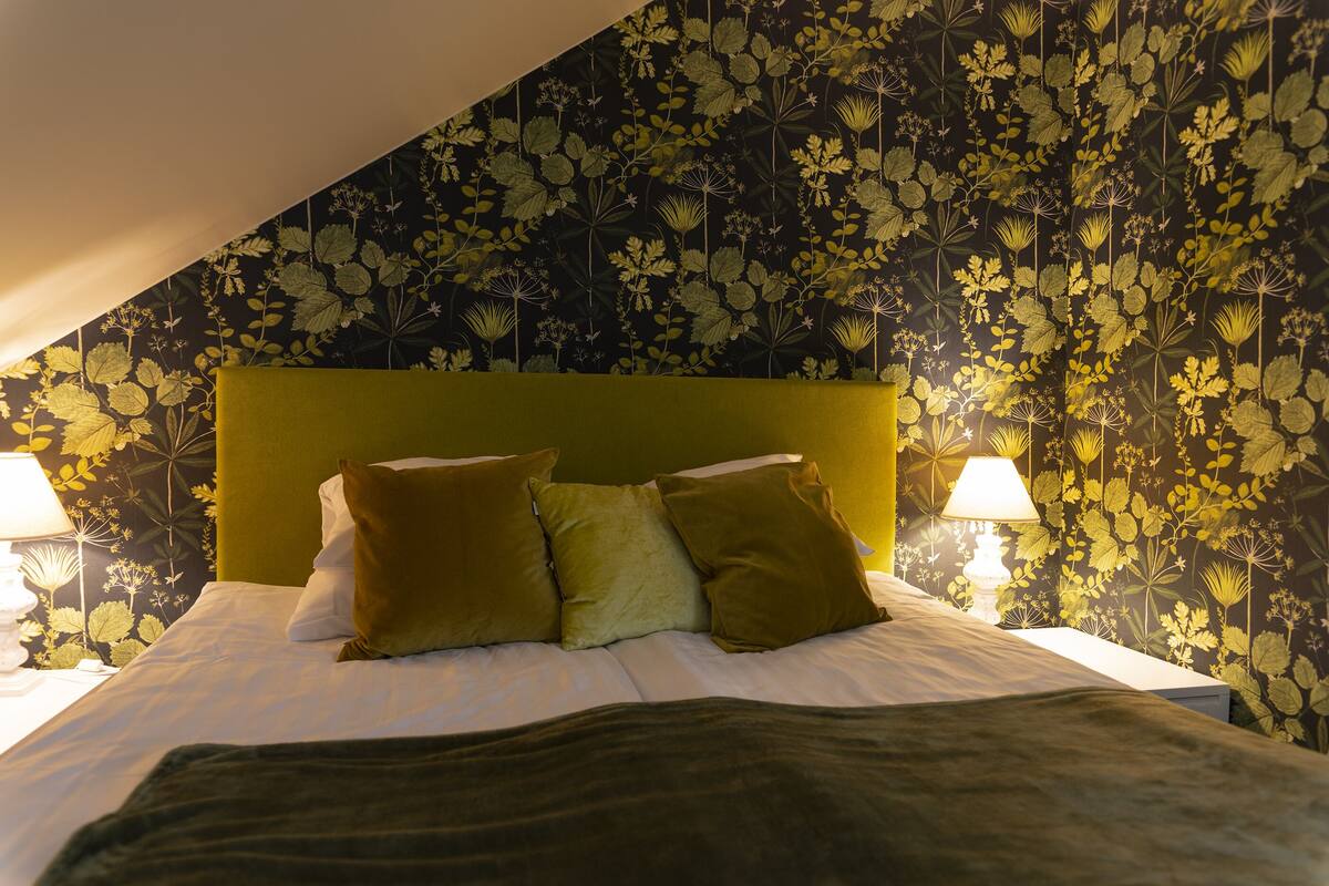 A bed with a green headboard, white bedding, and three pillows. There are two bedside lamps. The wall behind the bed has a floral wallpaper.