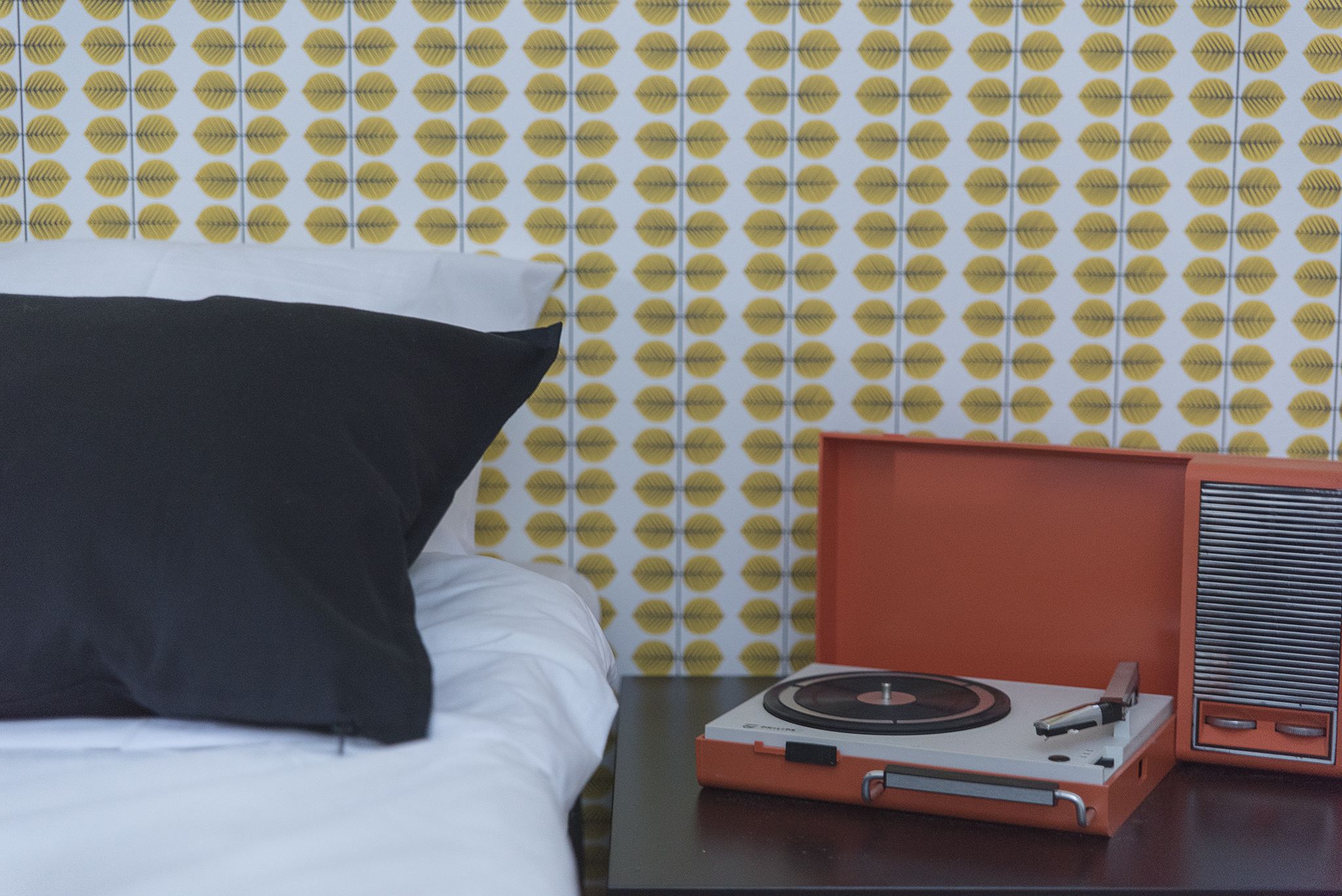 Una cama con un almohadón negro, una mesita pequeña con un tocadiscos y un papel tapiz estampado.