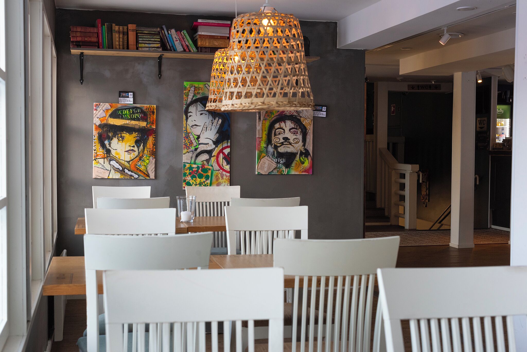 A dining area with white chairs and tables, a hanging woven light fixture, and artwork on the wall.