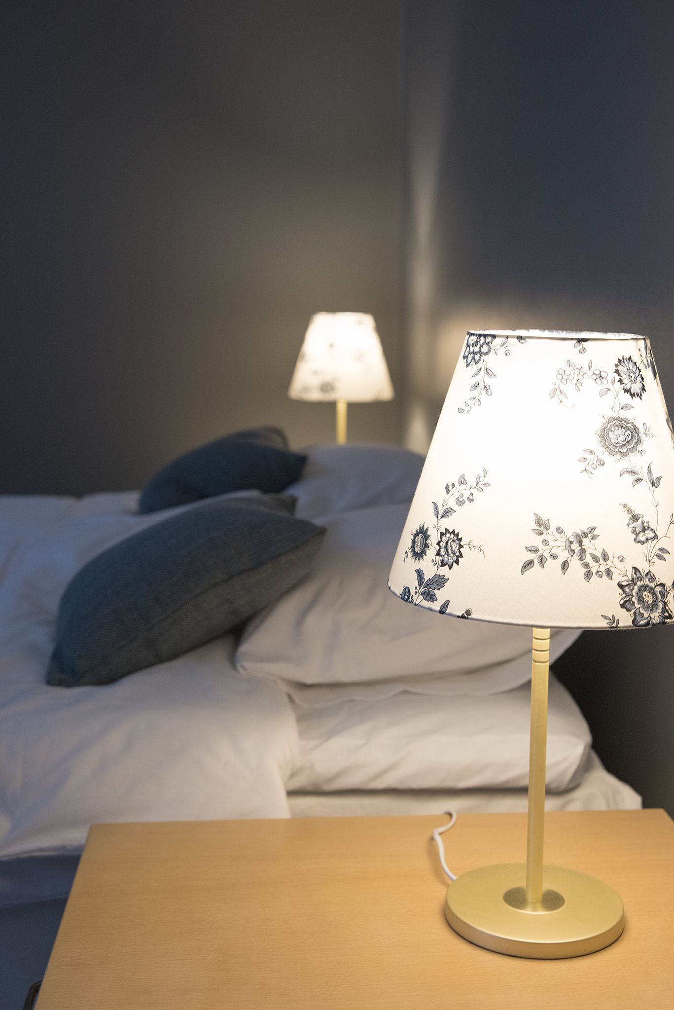 A bed with a white sheet and two pillows, a bedside table with a lamp, and a wall in the background.