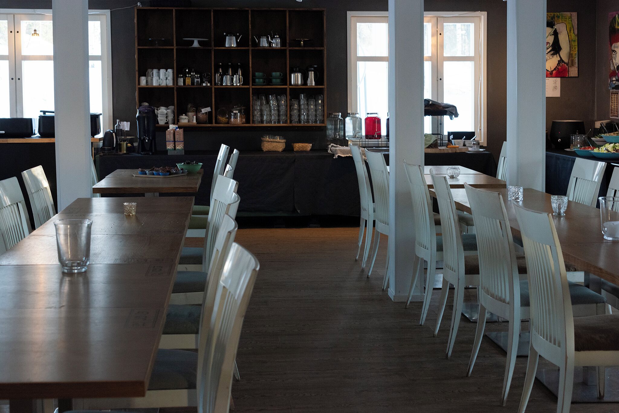 A dining area with a long wooden table and white chairs, a black cloth-covered table, and a bookshelf with various items.