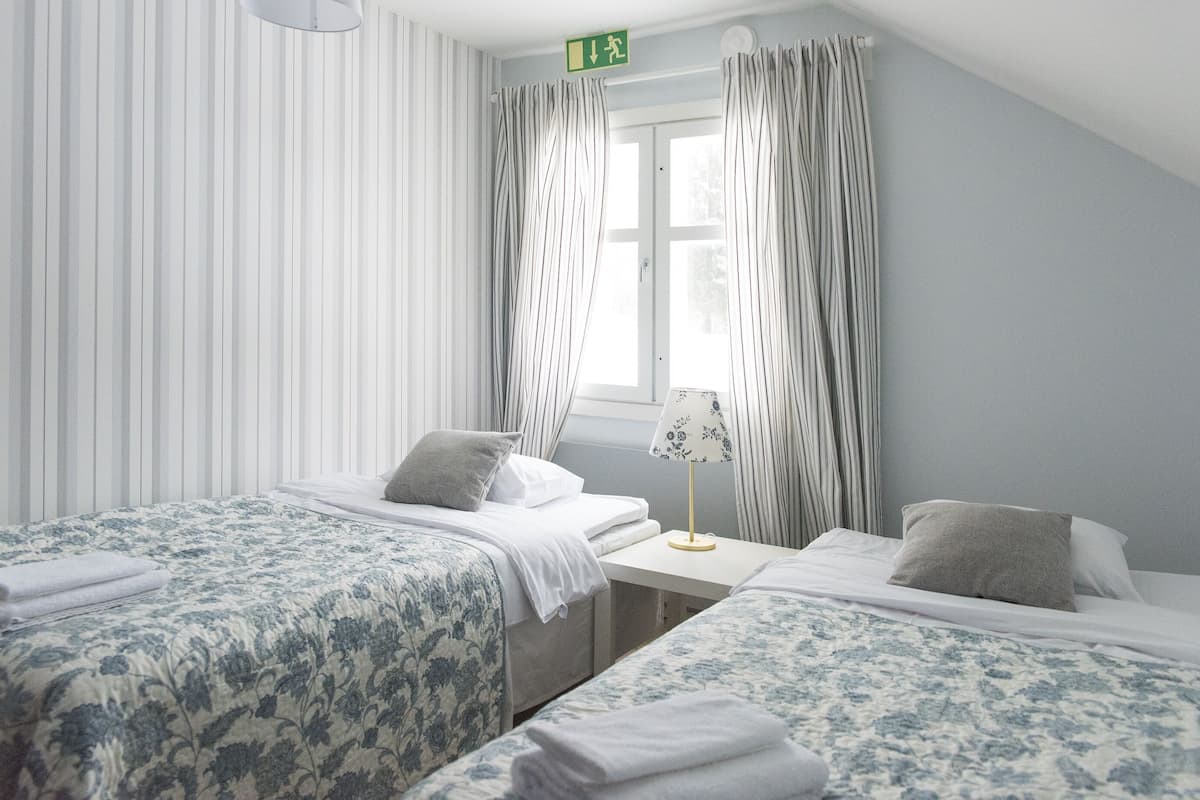 A hotel room with two beds, a small table, a lamp, and a window with curtains.