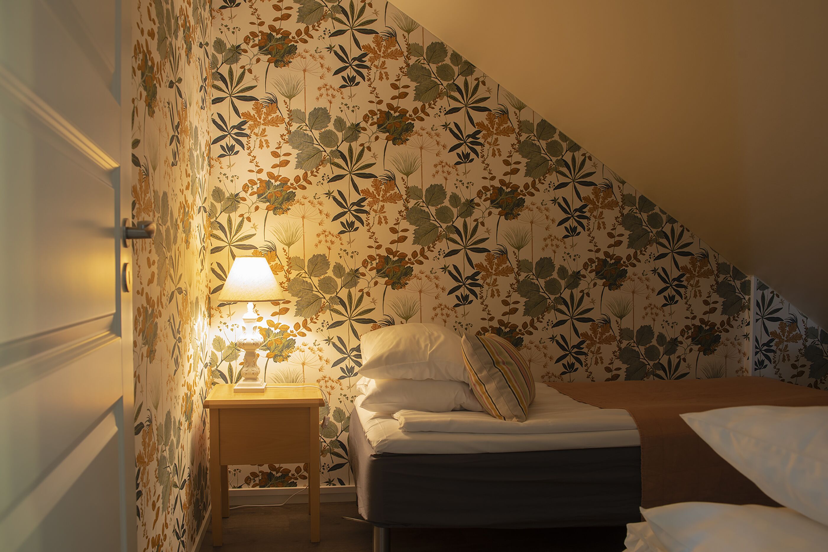Un dormitori amb paper pintat floral, una tauleta de nit amb una làmpada i un llit ben fet.