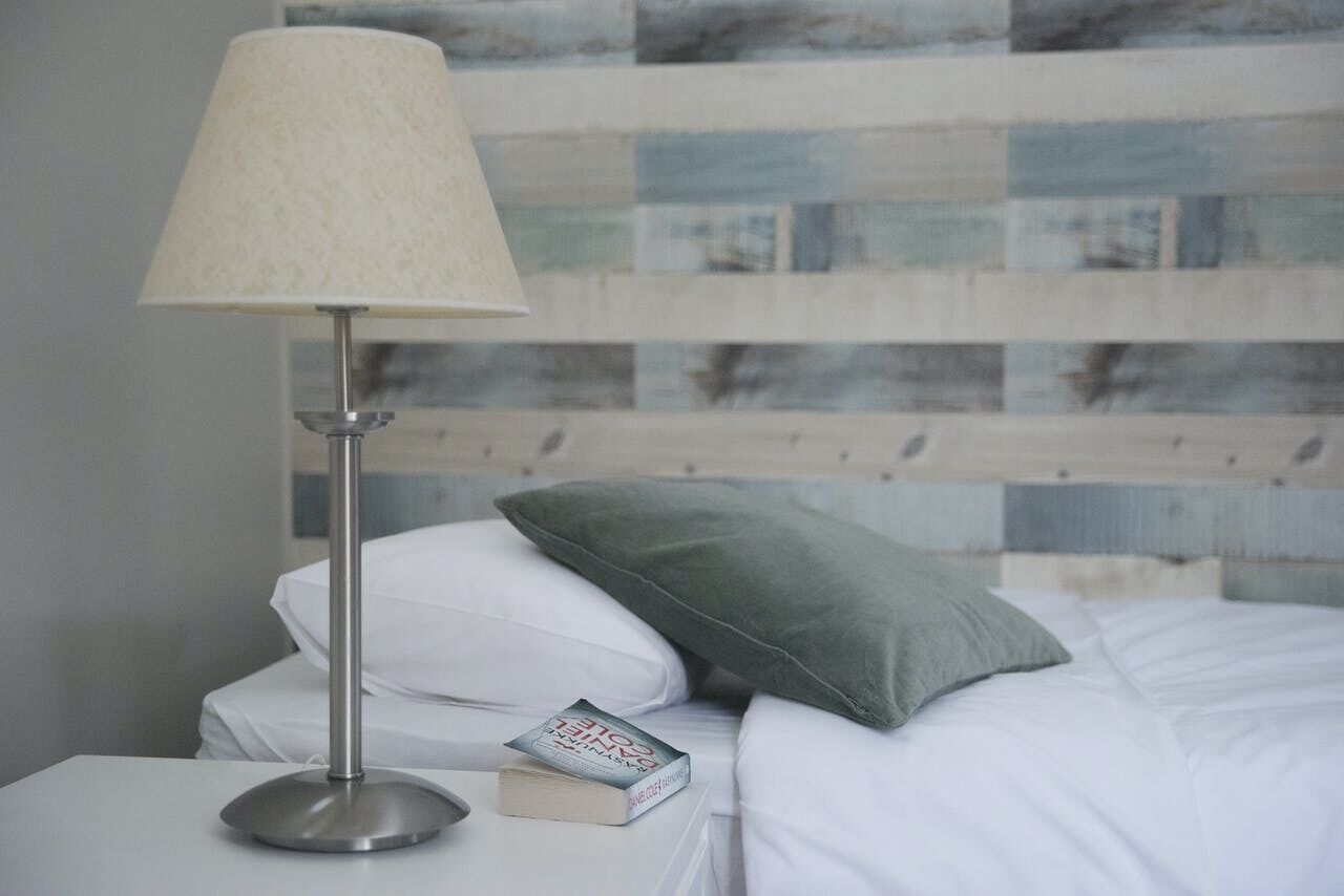 A neatly made bed with white linens, a green pillow, and a book titled "ONE OF A KIND" on a bedside table.