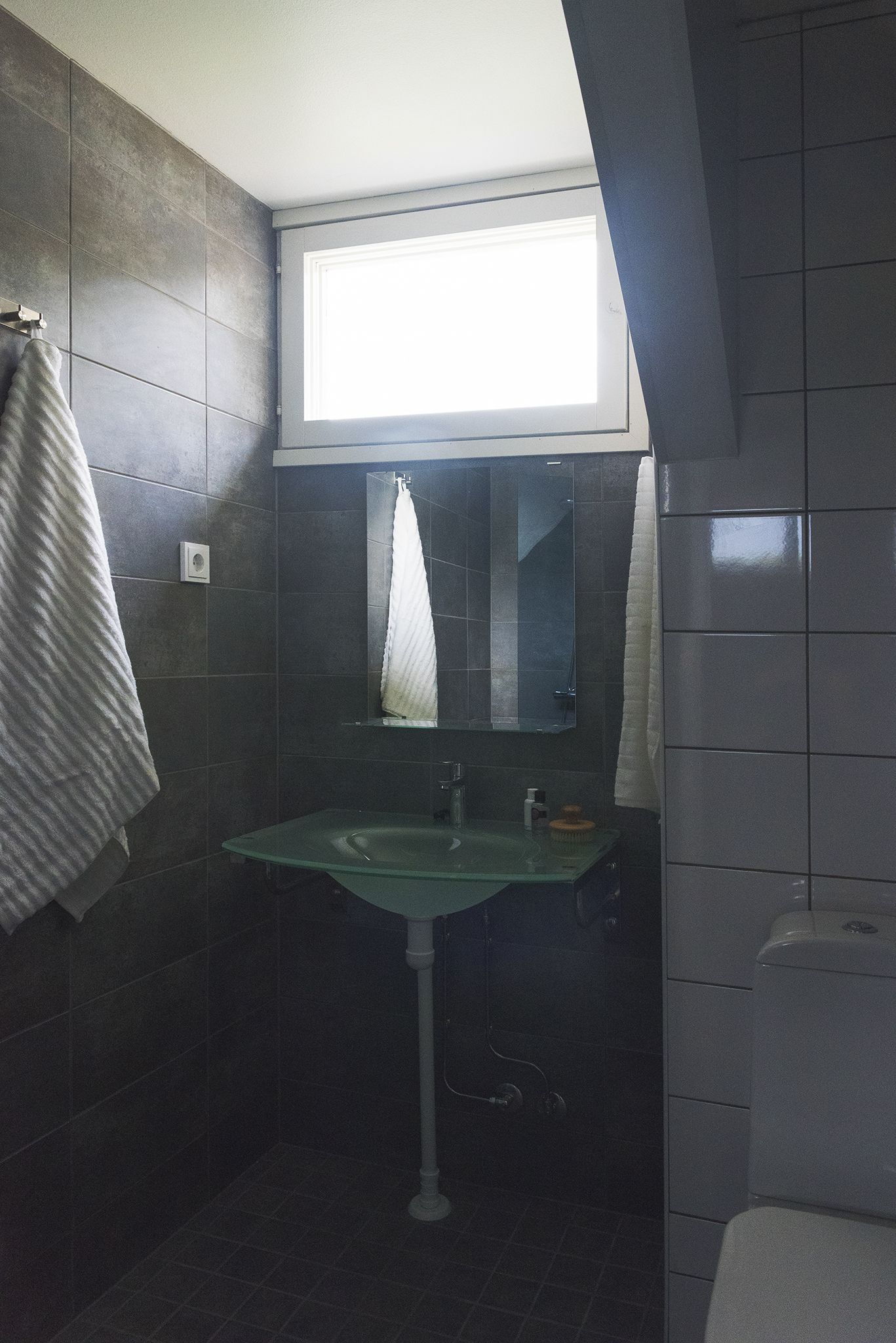 Un baño con baldosas oscuras, un lavamanos de vidrio, una ventana sobre el espejo y una toalla colgada en la pared.