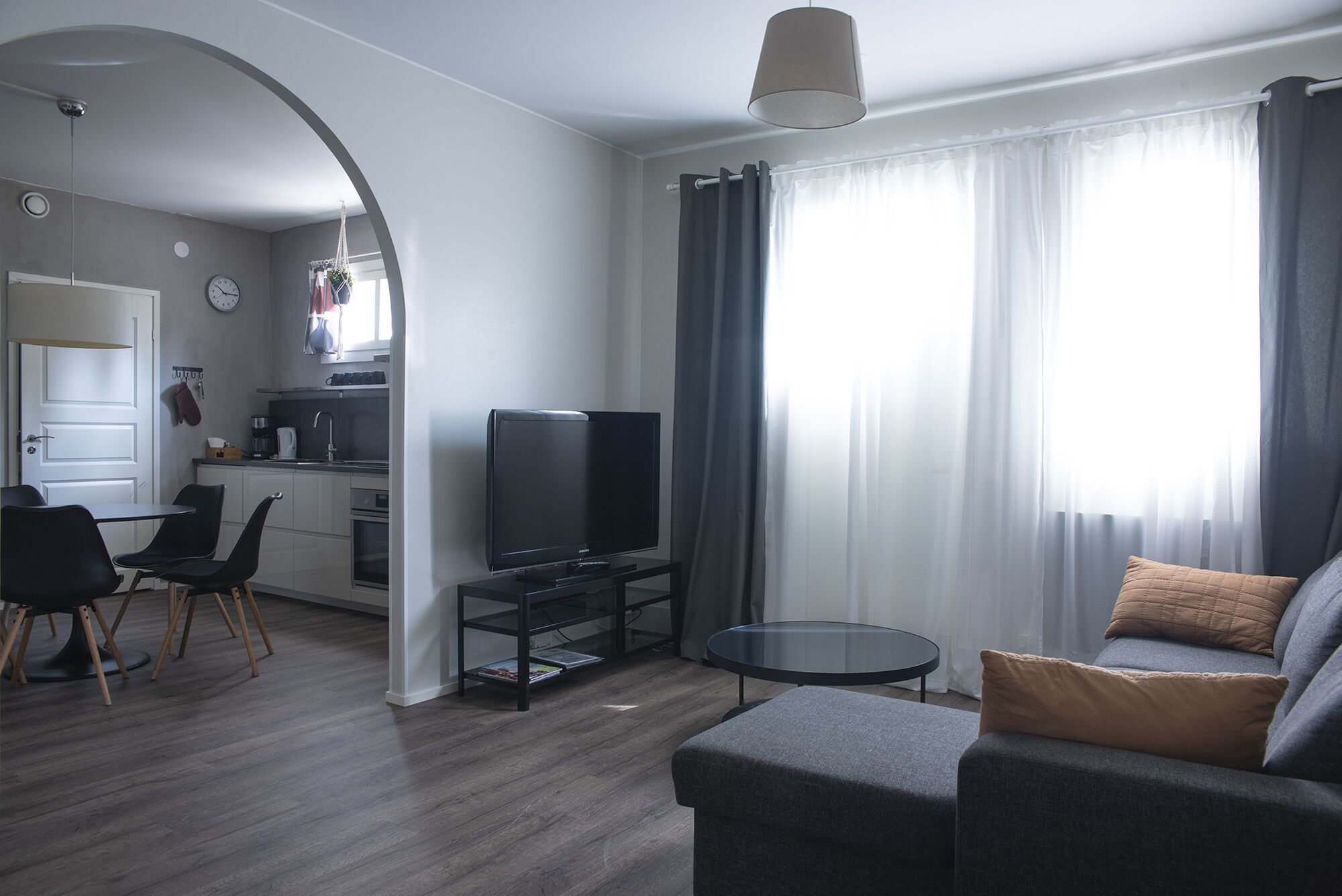 Un saló modern amb un sofà gris, una televisió i una taula baixa. Hi ha una zona de cuina amb un rentaplat i un rellotge a la paret.