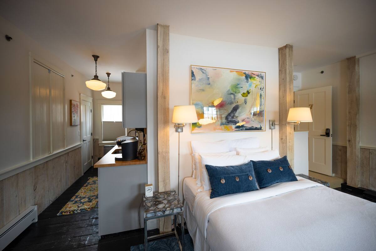 A hotel room with a bed, two bedside lamps, a painting on the wall, and a small table with a kettle.