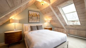 A bedroom with a sloped ceiling, a skylight, a bed with white bedding, two bedside tables with lamps, and a painting on the wall.