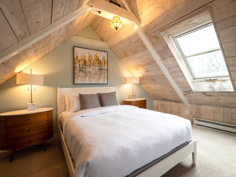 A bedroom with a sloped ceiling, a skylight, a bed with white bedding, two bedside tables with lamps, and a painting on the wall.