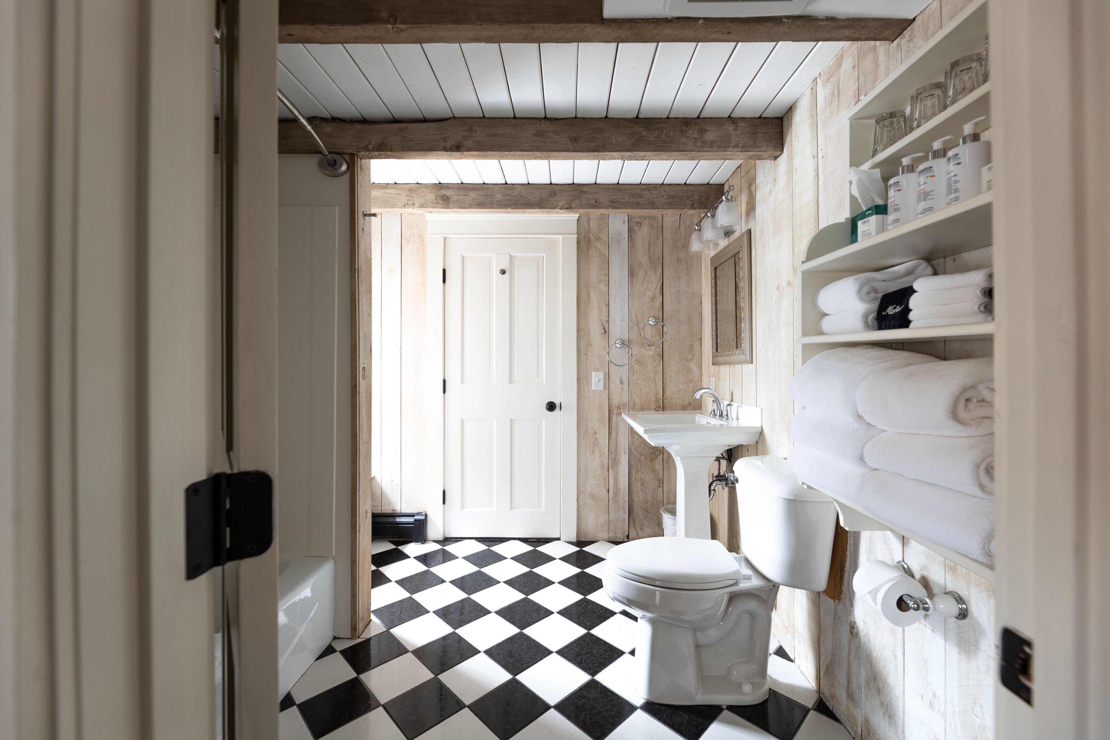 A bathroom with a black and white checkered floor, a white toilet, and a sink with a mirror above it. There are shelves with towels and toiletries.