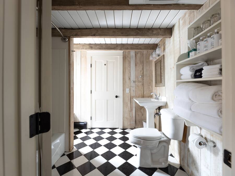 A bathroom with a black and white checkered floor, a white toilet, and a sink with a mirror above it. There are shelves with towels and toiletries.