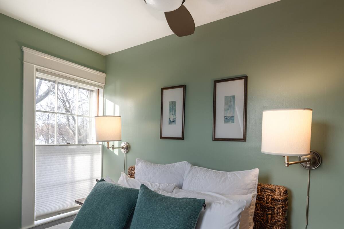 A bedroom with a bed, two pillows, a ceiling fan, two wall-mounted lamps, and two framed pictures.