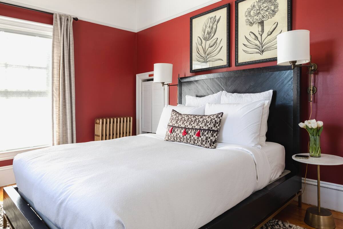 A neatly made bed with white linens and patterned pillows, a bedside table with a lamp, and a vase with flowers.